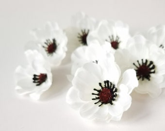 May include: A collection of artificial white anemone flowers, each featuring multiple rounded petals and a dark, textured center with deep red and black stamens. These small floral embellishments are arranged in a loose cluster.