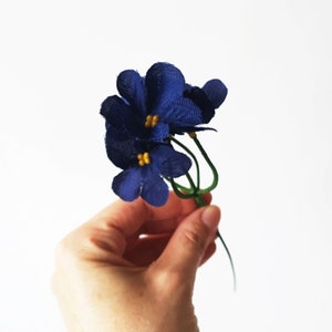 May include: A bunch of small blue fabric flowers with yellow centers. The flowers are on a green stem and are held in a hand.