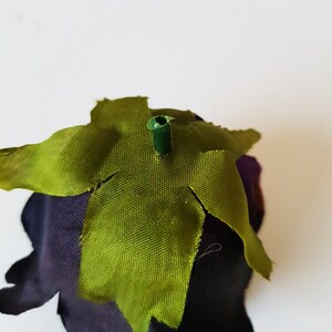 May include: A dark purple fabric flower head featuring a central green plastic stem protrusion surrounded by a star-shaped green fabric calyx with pointed edges. The petals are deep violet and have a smooth, matte texture throughout the piece.