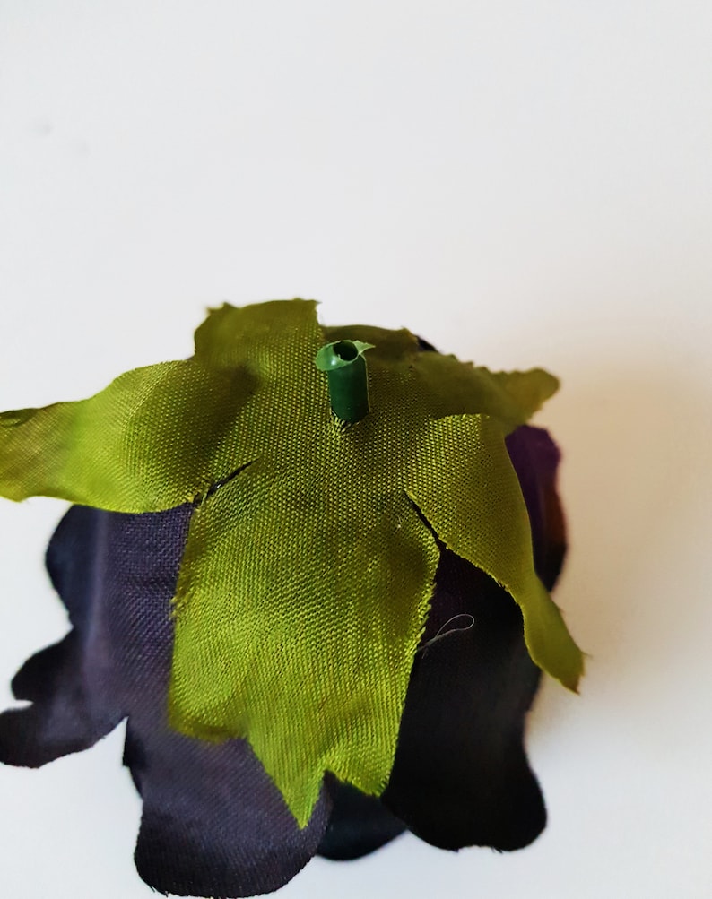 May include: A dark purple fabric flower head featuring a central green plastic stem protrusion surrounded by a star-shaped green fabric calyx with pointed edges. The petals are deep violet and have a smooth, matte texture throughout the piece.