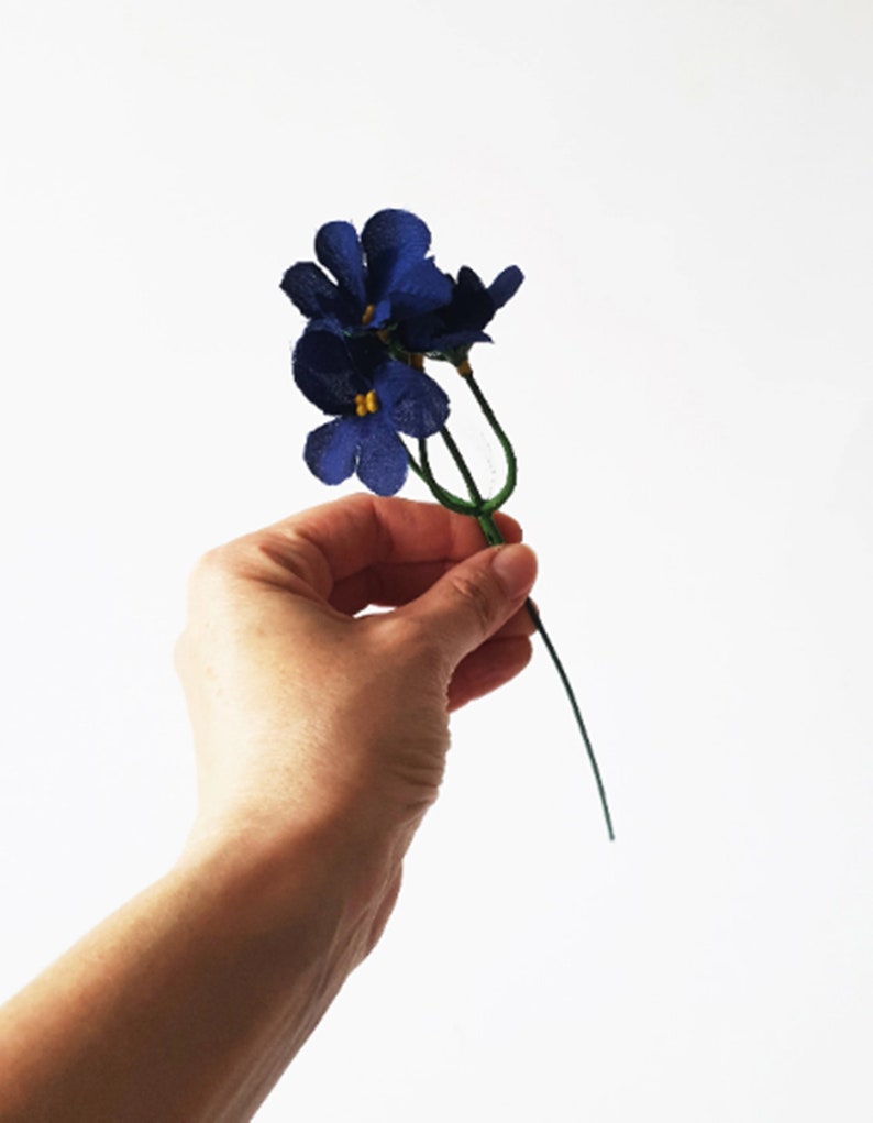 May include: A small bouquet of three blue felt flowers with yellow centers. The flowers are attached to a single green stem.