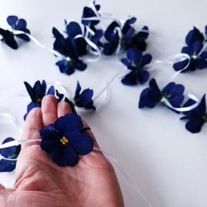Può includere: Una ghirlanda di fiori artificiali blu scuro con centri gialli, infilati su un sottile nastro bianco. I singoli fiori presentano petali arrotondati e sono distanziati uniformemente lungo la lunghezza del nastro bianco.