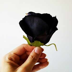 May include: A single artificial rose flower featuring deep black, fabric-textured petals that form a tight, rounded bloom. The flower includes a small, bright green plastic calyx at the base, providing a sharp color contrast to the dark petals.