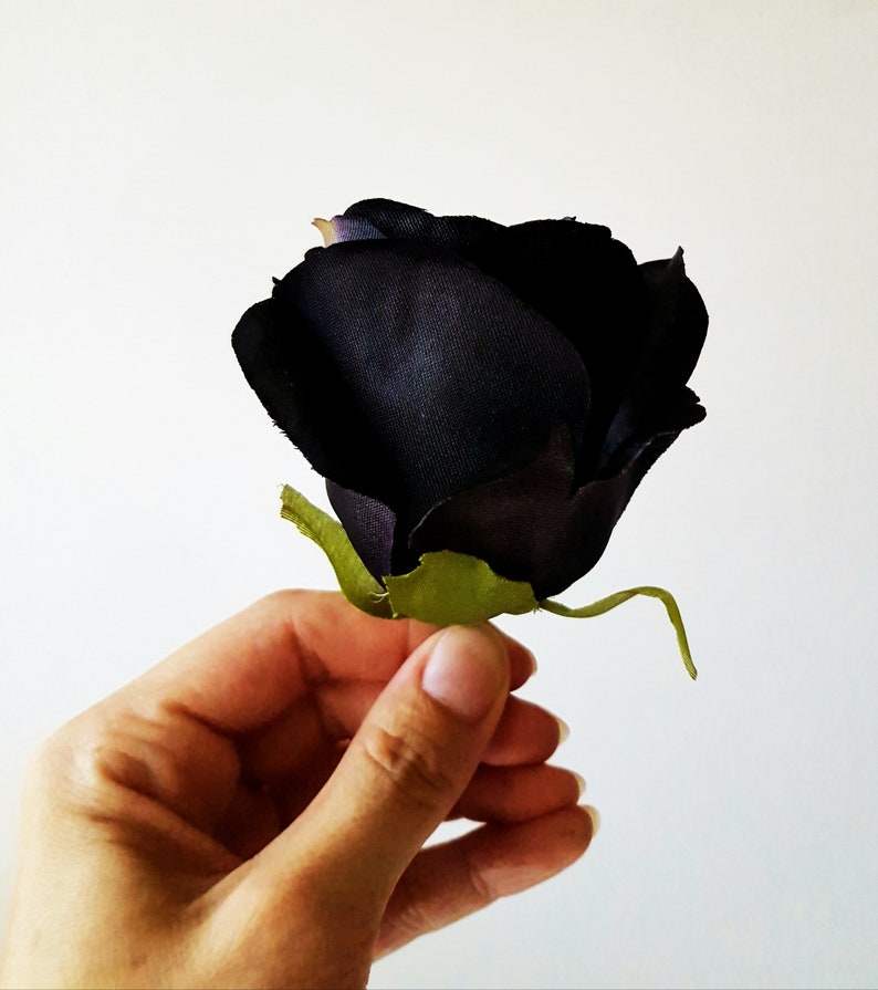 May include: A single artificial rose flower featuring deep black, fabric-textured petals that form a tight, rounded bloom. The flower includes a small, bright green plastic calyx at the base, providing a sharp color contrast to the dark petals.