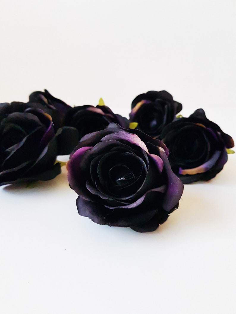 May include: A cluster of artificial rose heads in a deep black color with subtle hints of dark purple on the petal edges. Each flower features layered, rounded petals arranged in a classic bloom shape, with small green sepals visible at the base.