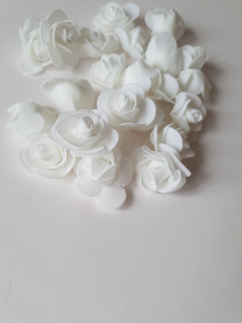 K&ouml;nnte beinhalten: Eine Ansammlung kleiner, einfarbig wei&szlig;er k&uuml;nstlicher Rosenbl&uuml;ten mit geschichteten Bl&uuml;tenbl&auml;ttern. Jede Blume hat eine abgerundete, bl&uuml;hende Form mit glatten, matten Oberfl&auml;chen, die in einer Gruppe angeordnet sind, um ihre einheitliche Struktur zu zeigen.