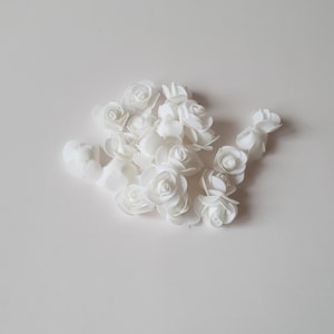K&ouml;nnte beinhalten: Eine Ansammlung kleiner, wei&szlig;er k&uuml;nstlicher Rosenbl&uuml;ten, die in einer Gruppe angeordnet sind. Jede einzelne Bl&uuml;te weist geschichtete, abgerundete Bl&uuml;tenbl&auml;tter auf, die ein enges, spiralf&ouml;rmiges Zentrum bilden und ein einheitliches Aussehen erzeugen.