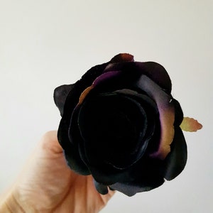 May include: A single artificial rose flower featuring layered petals in a deep black hue with subtle hints of dark purple and muted brown along the outer edges. The flower head is fully bloomed with a dense, rounded shape and soft fabric texture.