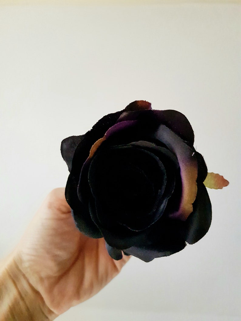 May include: A single artificial rose flower featuring layered petals in a deep black hue with subtle hints of dark purple and muted brown along the outer edges. The flower head is fully bloomed with a dense, rounded shape and soft fabric texture.