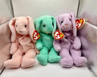 Può includere: Tre coniglietti di peluche dai colori pastello: rosa, verde menta e lavanda. Ogni coniglietto ha lunghe orecchie, un nastro e un'etichetta a forma di cuore. I coniglietti sono seduti uno accanto all'altro.