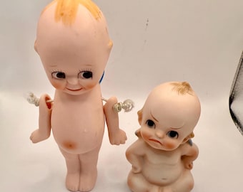 Può includere: Due bambole Kewpie antiche. Una bambola è in piedi con le braccia staccate, e l'altra è seduta con un'espressione arrabbiata. Entrambe le bambole hanno tratti del viso dipinti, tra cui occhi scuri e guance rosee. Le bambole sono fatte di porcellana.