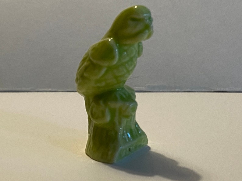 Pu&ograve; includere: Una statuetta di ceramica verde di un pappagallo appollaiato su un ceppo d'albero. Il pappagallo &egrave; rivolto a sinistra e ha la testa inclinata verso il basso.