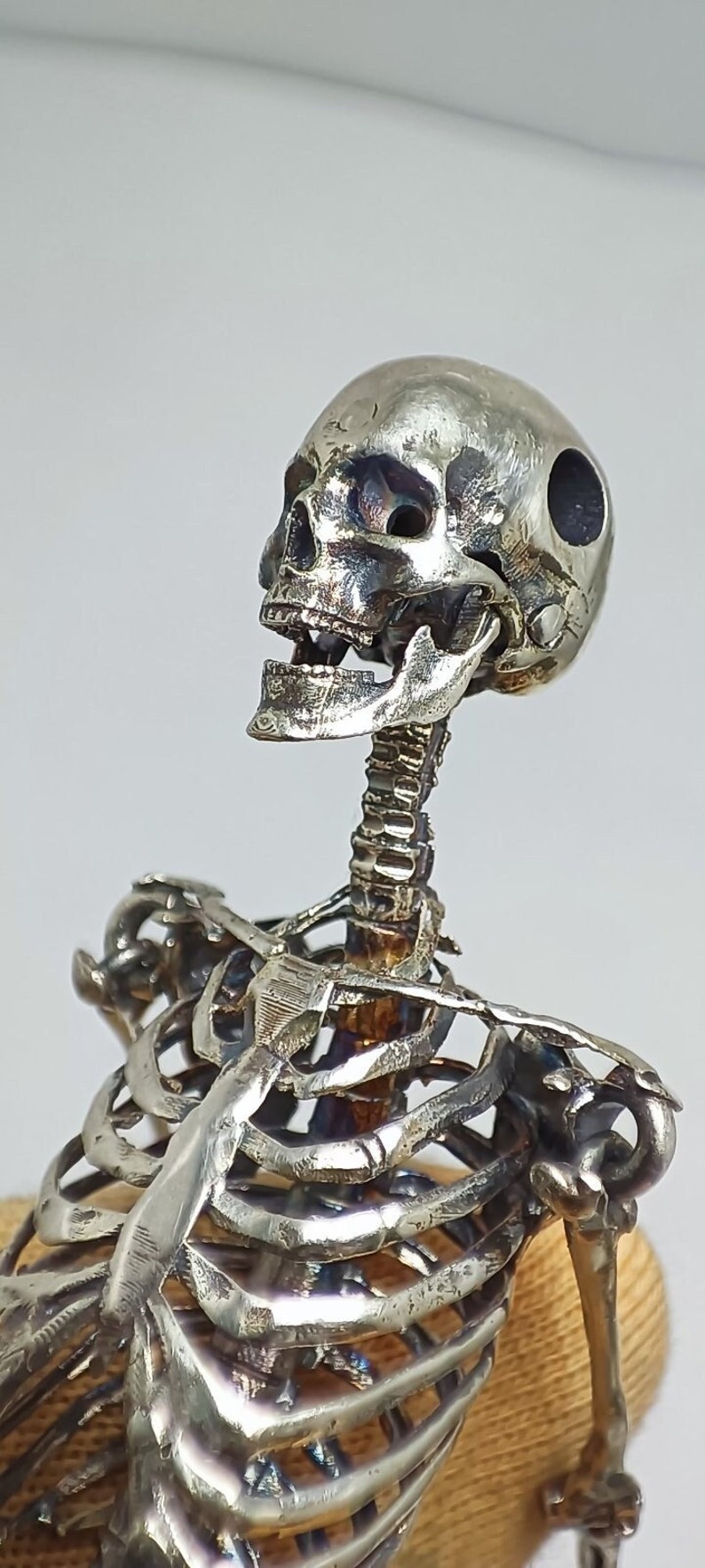 Puede incluir: Una escultura de esqueleto de plata con un cr&aacute;neo y una caja tor&aacute;cica detallados. El esqueleto est&aacute; en posici&oacute;n sentada con la cabeza inclinada hacia atr&aacute;s.
