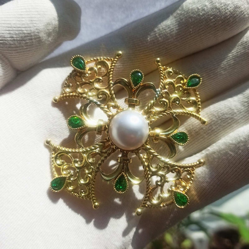 May include: A gold filigree brooch with a large white pearl in the center and green gemstones around the edges.