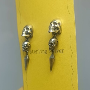 May include: A pair of sterling silver earrings featuring two skull charms dangling from a pointed spike. The earrings are displayed on a yellow card with the text "sterling silver".
