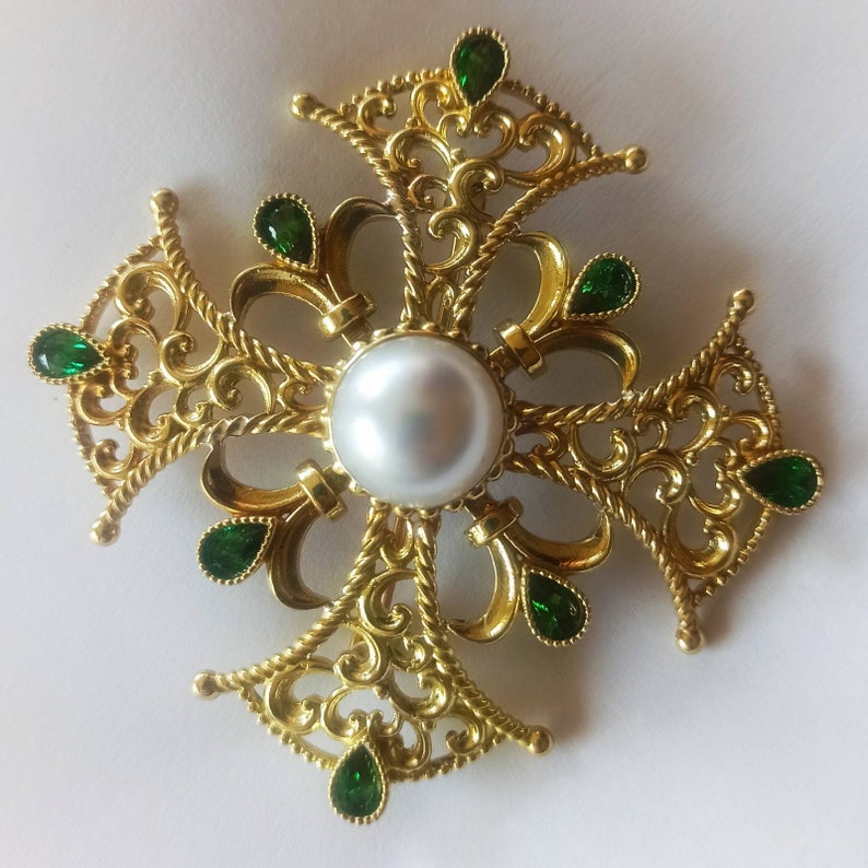 May include: A gold filigree cross brooch with a white pearl in the center and green gemstones around the edges.