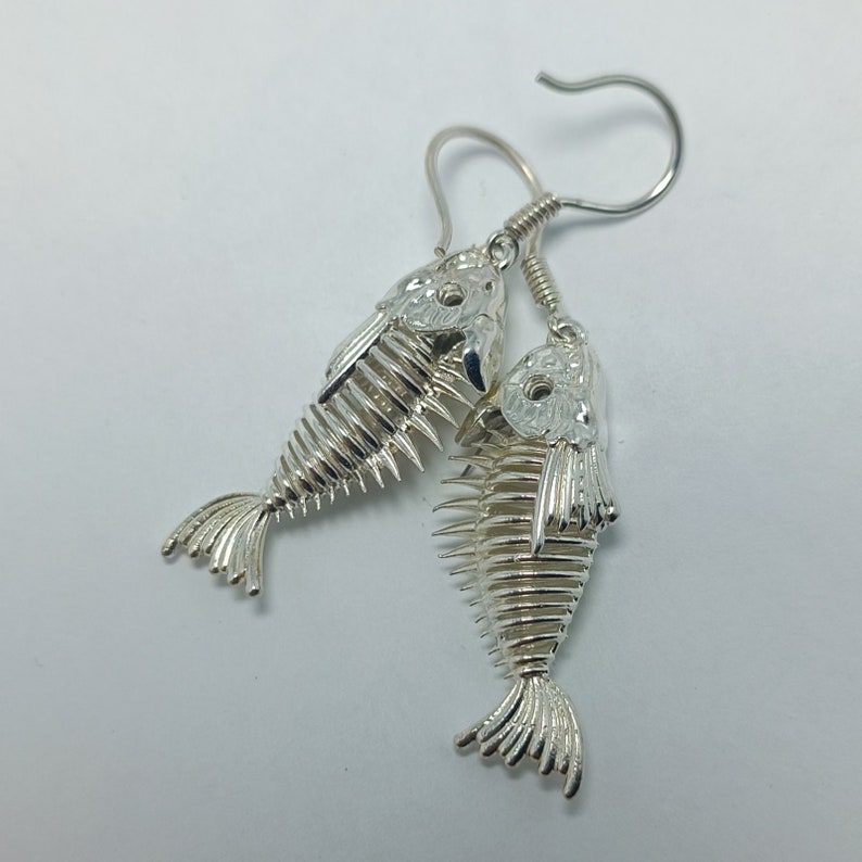 May include: Silver fish skeleton earrings with a detailed design. The earrings are dangling and have a hook closure.