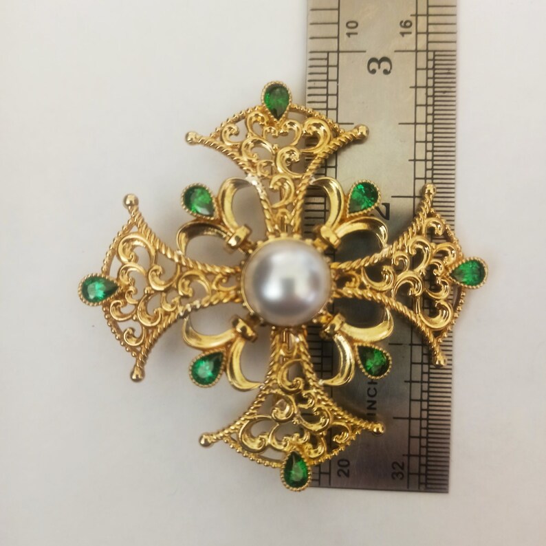 May include: A gold filigree cross with a white pearl in the center and green gemstones around the edges. The cross is about 3 inches wide.