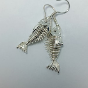 May include: Silver fish skeleton earrings with a detailed design. The earrings are hanging from a silver hook.