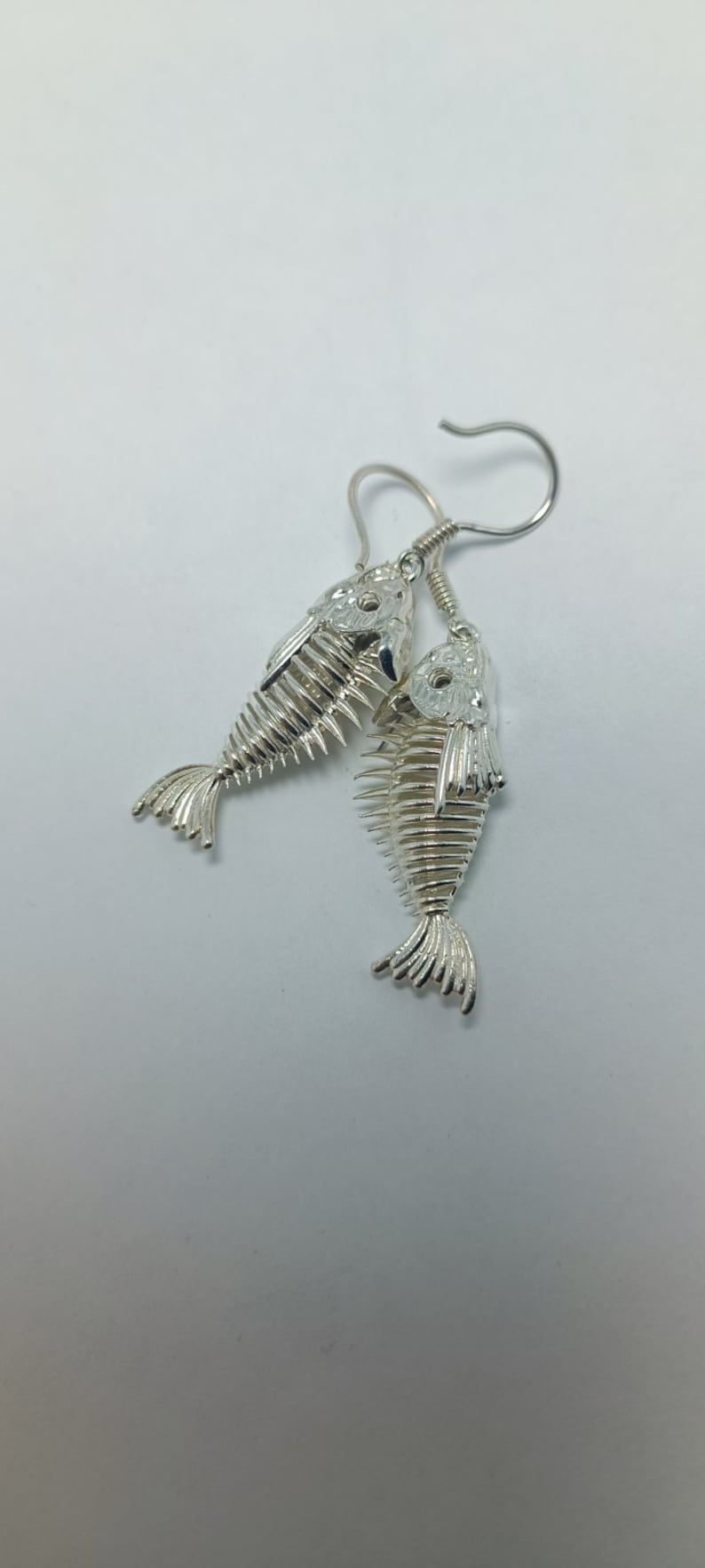 May include: Silver fish skeleton earrings with a detailed design. The earrings are hanging from a silver hook.