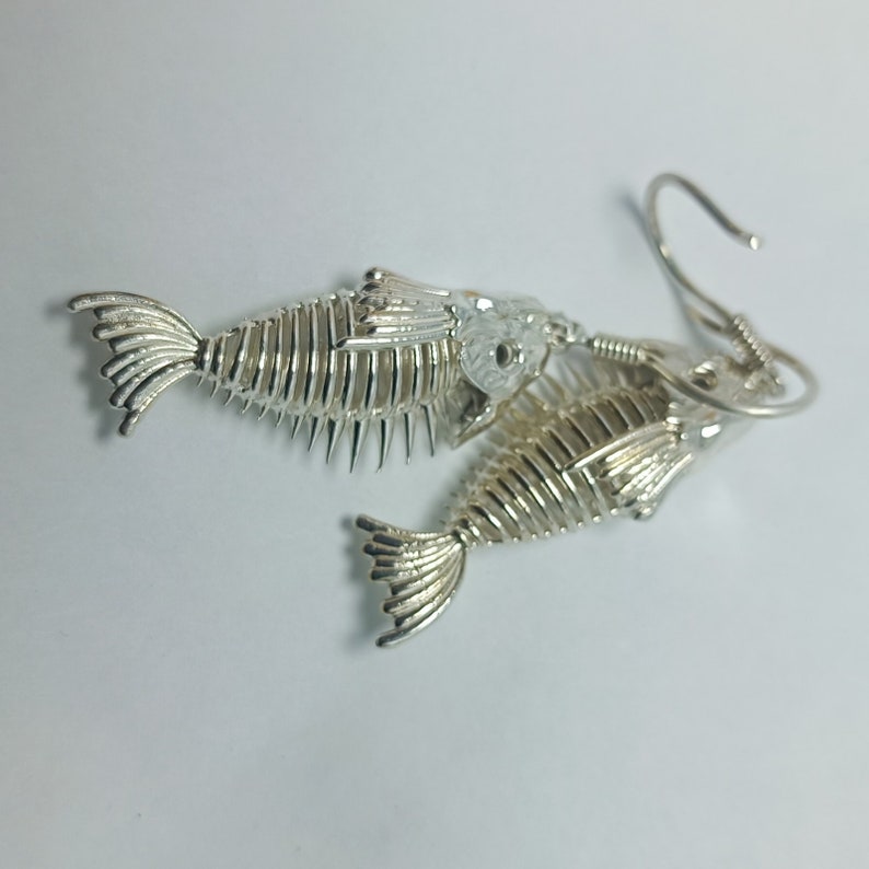 May include: Silver fish skeleton earrings with a detailed design. The earrings are dangling and have a hook closure.