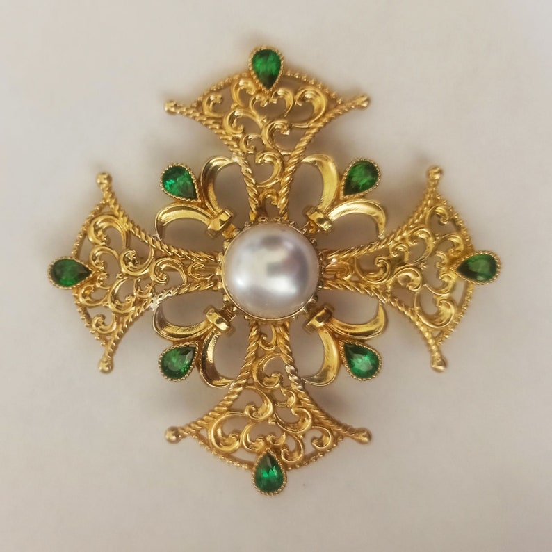 May include: A gold filigree cross pendant with a large white pearl in the center and green gemstones around the edges.