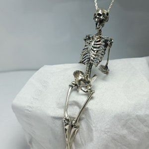 Puede incluir: Un collar con un colgante de esqueleto de plata. El esqueleto est&aacute; sentado con las piernas cruzadas y los brazos apoyados en las rodillas. El collar est&aacute; hecho de una cadena de plata delicada.