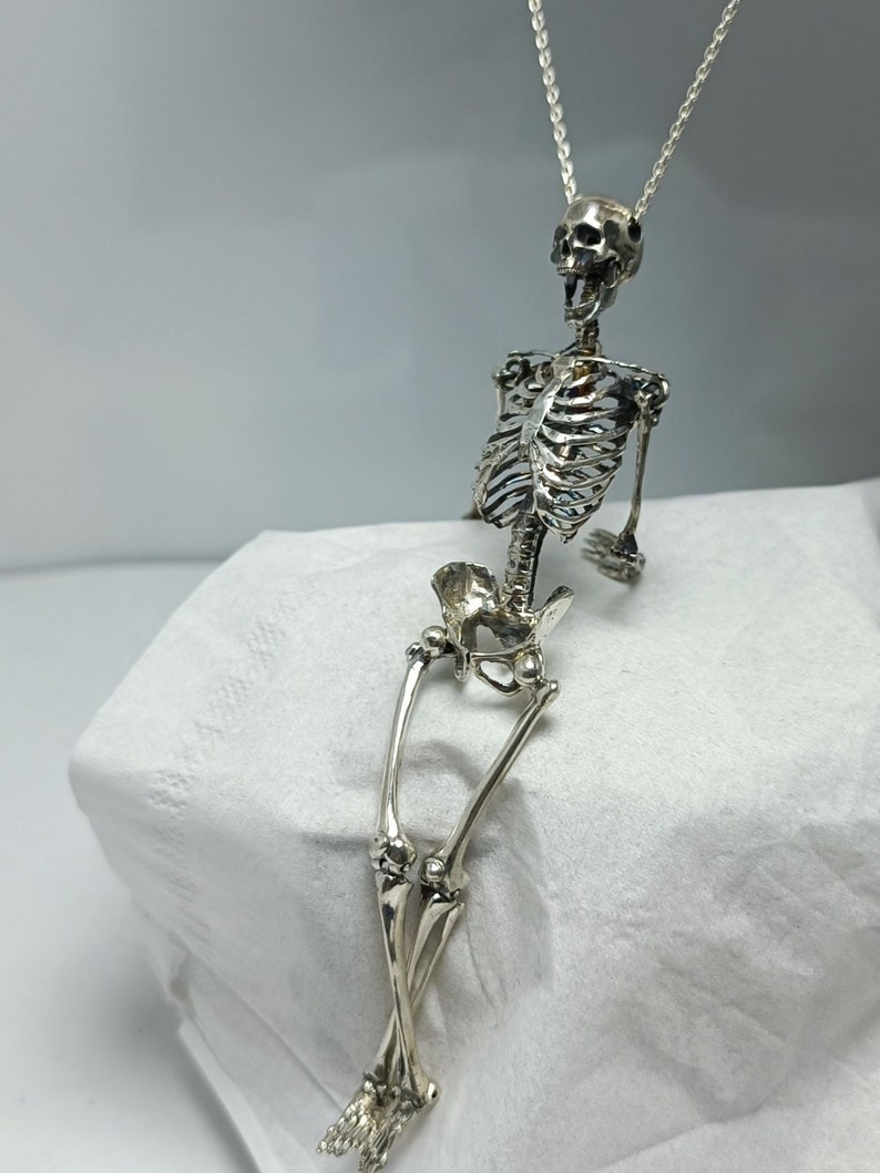 Puede incluir: Un collar con un colgante de esqueleto de plata. El esqueleto est&aacute; sentado con las piernas cruzadas y los brazos apoyados en las rodillas. El collar est&aacute; hecho de una cadena de plata delicada.