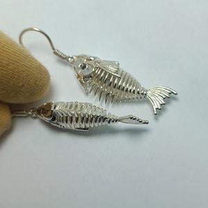 May include: A pair of silver fish skeleton earrings. The earrings are made of metal and have a detailed design of a fish skeleton. The earrings are hanging from a silver hook.