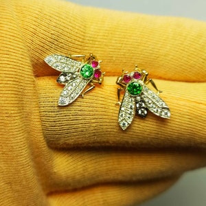 May include: A pair of gold bee earrings featuring emerald green and ruby red gemstones. The bees are decorated with clear sparkling stones.