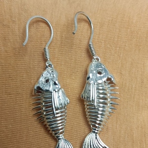 May include: A pair of silver fish skeleton earrings with a dangling design. The earrings are made of metal and have a detailed fish skeleton design.