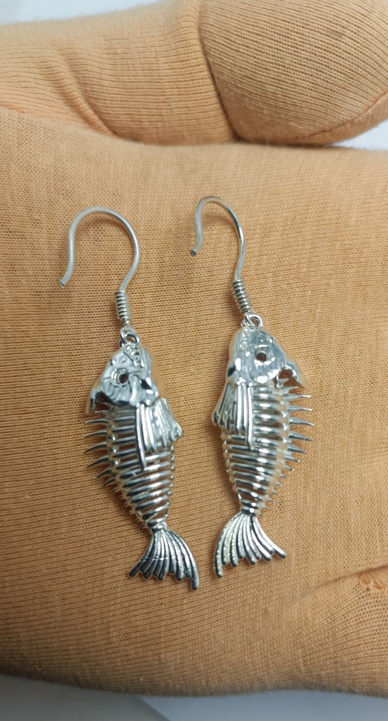 May include: A pair of silver fish skeleton earrings with a dangling design. The earrings are made of metal and have a detailed fish skeleton design.
