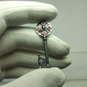 May include: A silver key with a skull design and pink gemstones. The key has a decorative axe-shaped handle.