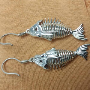 May include: A pair of silver fish skeleton earrings. The earrings are made of metal and have a detailed fish skeleton design. The earrings are hanging from silver hooks.