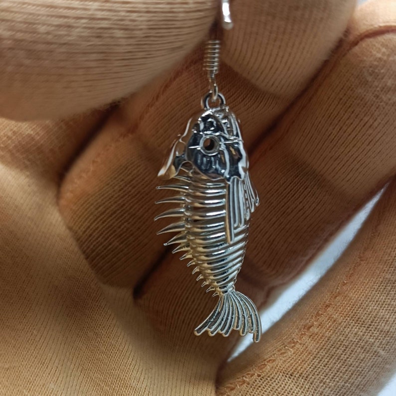 May include: Silver fish skeleton dangle earrings. The earrings are shaped like a fish skeleton with a detailed design.