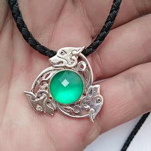 Pode incluir: Um pingente de nó celta de prata com uma gema verde no centro. O pingente está em um cordão trançado preto.
