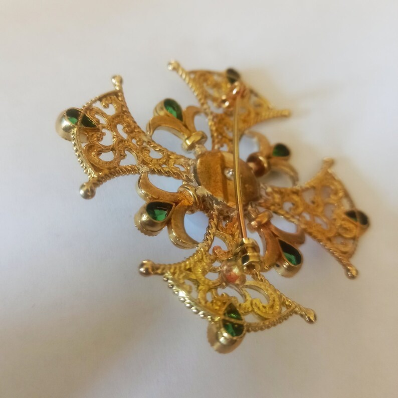 May include: A gold filigree brooch with green gemstones. The brooch is in the shape of a cross with a decorative design.