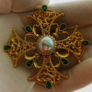 May include: A gold filigree cross pendant with a large white pearl in the center and green gemstones around the edges.
