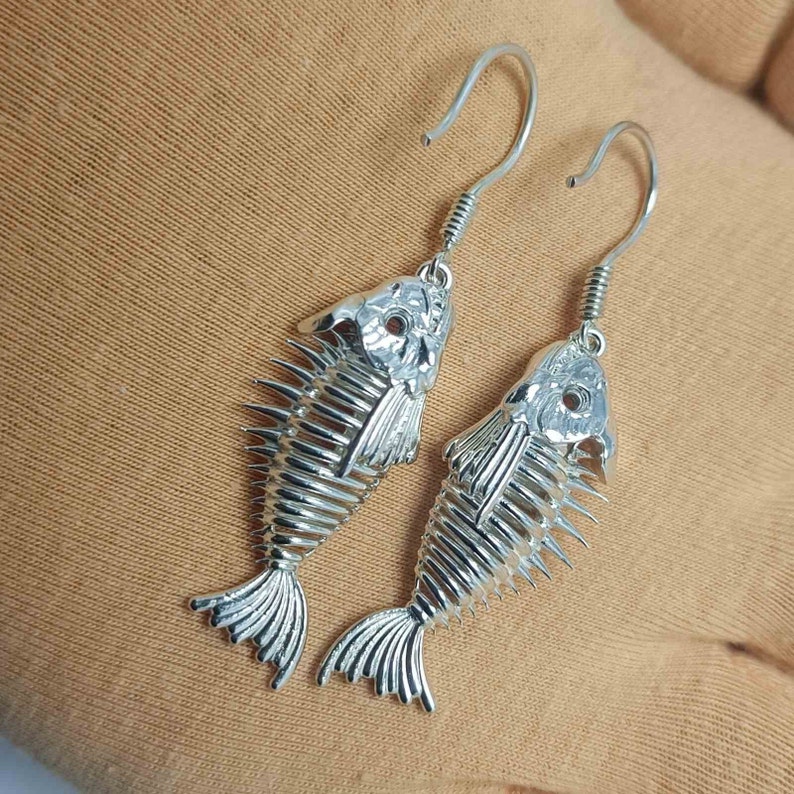 May include: A pair of silver fish skeleton earrings with a hook closure. The earrings are detailed and have a realistic fish skeleton design.