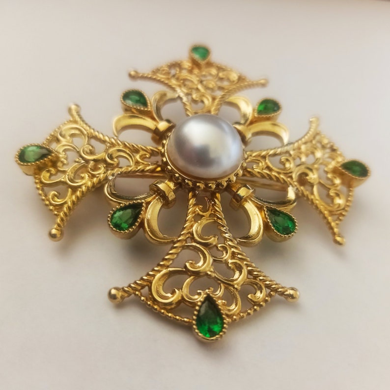 May include: A gold filigree cross brooch with a large white pearl in the center and green gemstones around the edges.