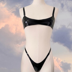 Puede incluir: Conjunto de lencería de látex negro, compuesto por un sujetador con tirantes ajustables y un tanga de corte alto. Las prendas brillantes y ajustadas se exhiben en un maniquí sobre un fondo de cielo nublado.