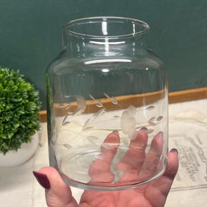 May include: Clear glass vase with etched floral and leaf design. The vase has a wide, cylindrical shape with a slightly flared rim. The etched design features delicate flowers and leaves, adding a touch of elegance. A decorative piece for home decor.