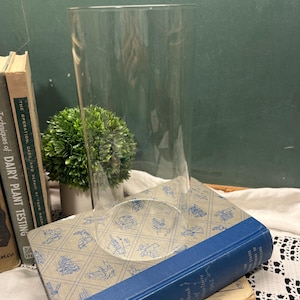 May include: A clear, cylindrical glass vase with an open top and a flat base. The vase is positioned upright on top of a stack of vintage hardcover books, including one titled The Yearbook of Agriculture 1962 and Dairy Plant Testing.