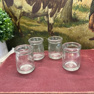 May include: Four small, clear glass jars with rounded bodies and flared rims are arranged on a red surface. The jars are empty and appear to be vintage. A green plant and a painting are in the background.