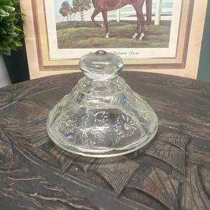 Peut inclure: Un couvercle en verre transparent avec un dessus arrondi et un motif floral. Le couvercle est posé sur une surface en bois sombre. Une image encadrée d'un cheval est en arrière-plan.