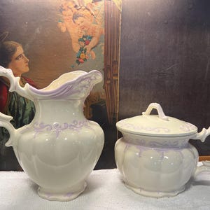 May include: A cream-colored ceramic pitcher and a matching lidded sugar bowl, both featuring ornate, raised lavender scrollwork details and scalloped edges. The pitcher has a curved handle and a pouring spout, while the bowl has a lid with a handle.
