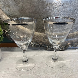 Peut inclure: Deux verres à vin transparents avec des bords argentés. Les verres ont un design classique avec une base à pied et un large bol. Les verres sont présentés sur une surface blanche, avec un plateau argenté décoratif en arrière-plan.