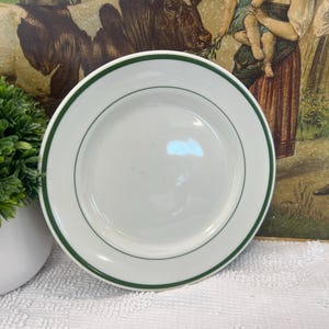 May include: A white ceramic plate with a green border. The plate is round and has a simple, classic design. The plate is likely for serving food. The plate is on a white surface.