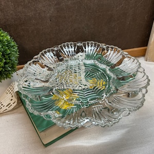 May include: A clear, scalloped-edge glass serving dish with a central compartment and radiating sections. The dish is resting on a green book and a white cloth, with a small green plant in the background. The dish has a decorative, textured design.