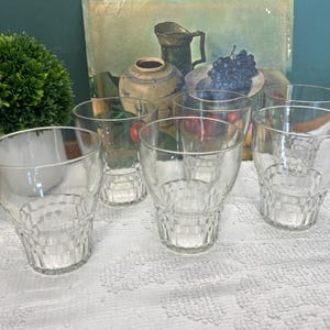 May include: Six clear drinking glasses with a textured, geometric pattern on the lower half. The glasses are arranged on a white, textured surface. A green plant and a still-life painting are in the background.